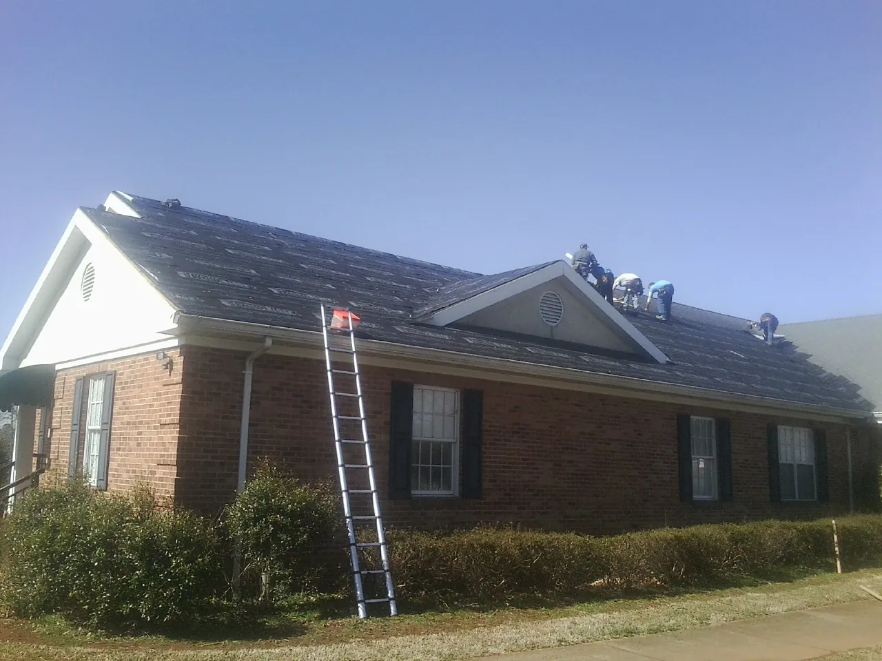 Crew tearing shingles off a brick ranch roof with a ladder set — Metal Roof Repair work by our crew in Bartonsville