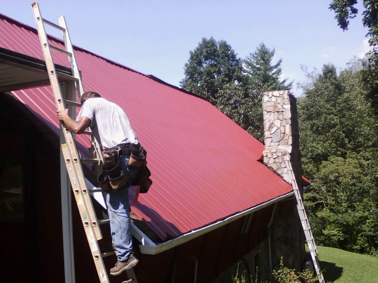 Roofer on a ladder beside a red metal roof and stone chimney — Soffit & Fascia Repair by Roofing Brothers in Bartonsville